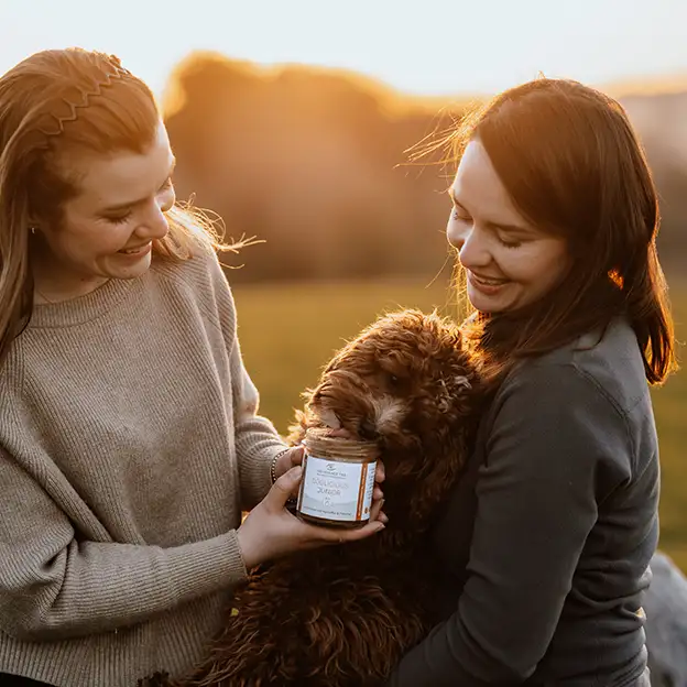 Zwei Tierhalterinnen halten einen Hund, der neugierig an einem Produktglas von „Das Gesunde Tier“ schnuppert.
