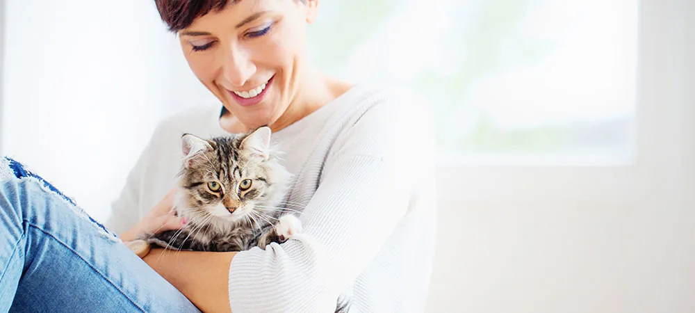 Lächelnde, auf dem Boden sitzende Frau mit kurzen Haaren hält kleine getigerte Katze im Arm.