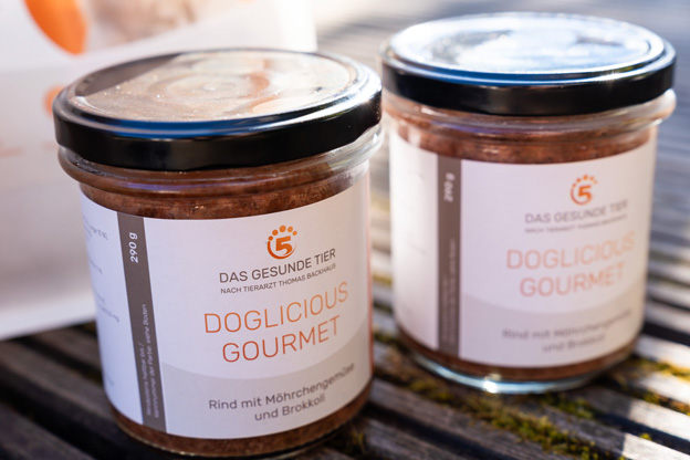Zwei Gläser Doglicious Gourmet Hundefutter von Das Gesunde Tier mit Rind, Möhrengrün und Brokkoli stehen auf einem Holztisch im Freien, aufgenommen bei warmem Sonnenlicht.
