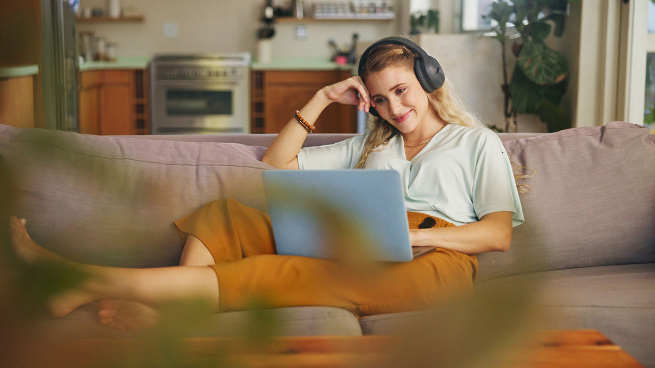 Eine junge Frau sitzt auf ihrer Couch am Laptop und lächelt.