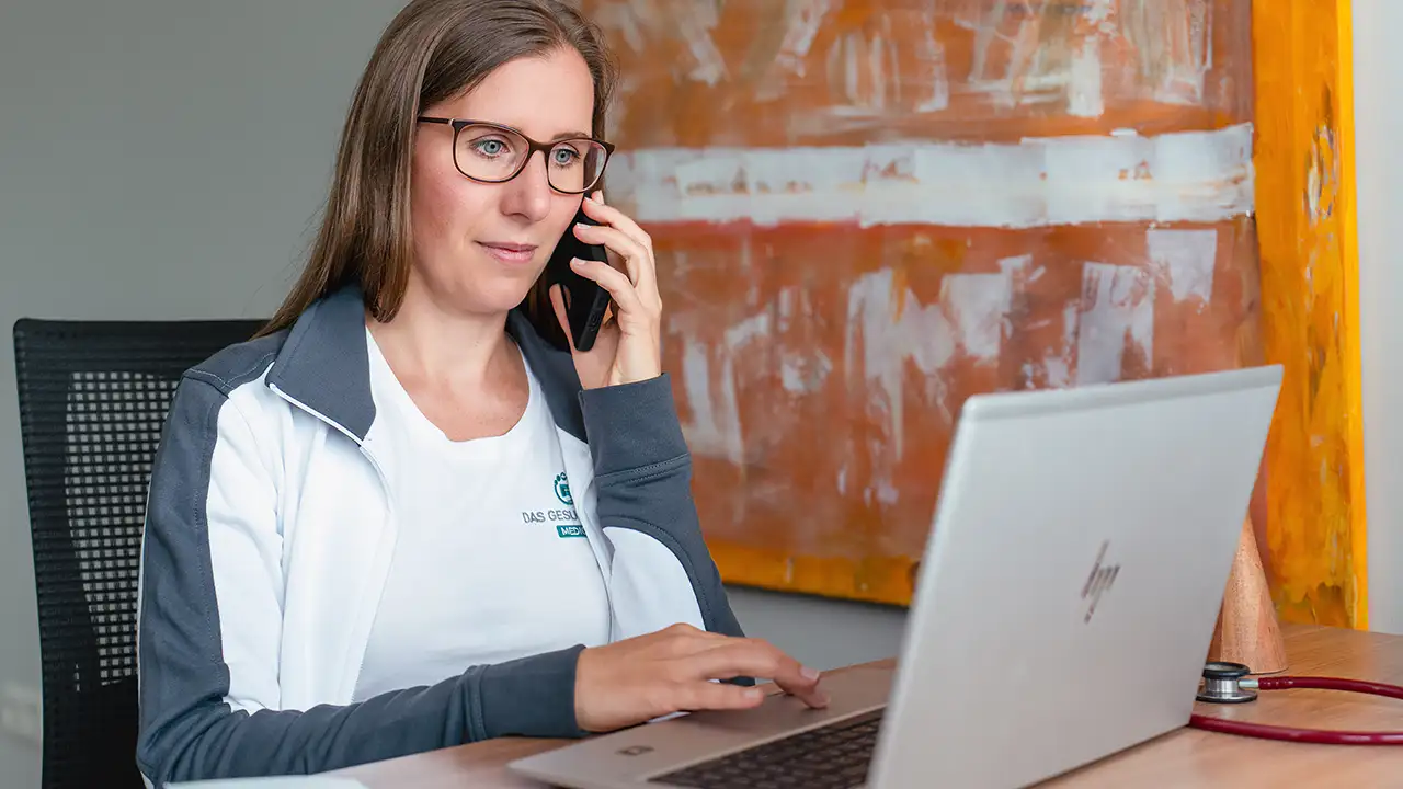 Eine Frau mit Brille, die am Schreibtisch am Laptop arbeitet und dabei telefoniert.