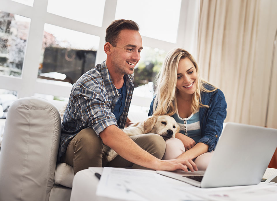 Eine Frau und ein Mann sitzen auf der Couch, gemeinsam mit ihrem Hund und schauen auf einen Laptop.