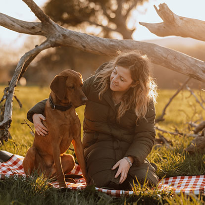 Frau sitzt mit Hund auf einer Picknickdecke unter einem Baumstamm im Sonnenuntergang.