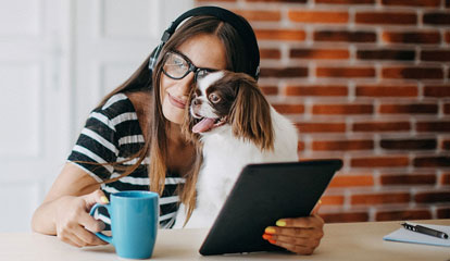 Eine Frau mit Kopfhöhrern hält ihr Tablet und eine Tasse Kaffee und kuschelt dabei mit ihrem Hund.