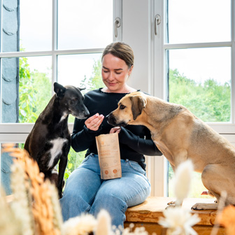 Frau füttert zwei Hunde mit Snacks von Das Gesunde Tier aus einer braunen Snack-Packung.